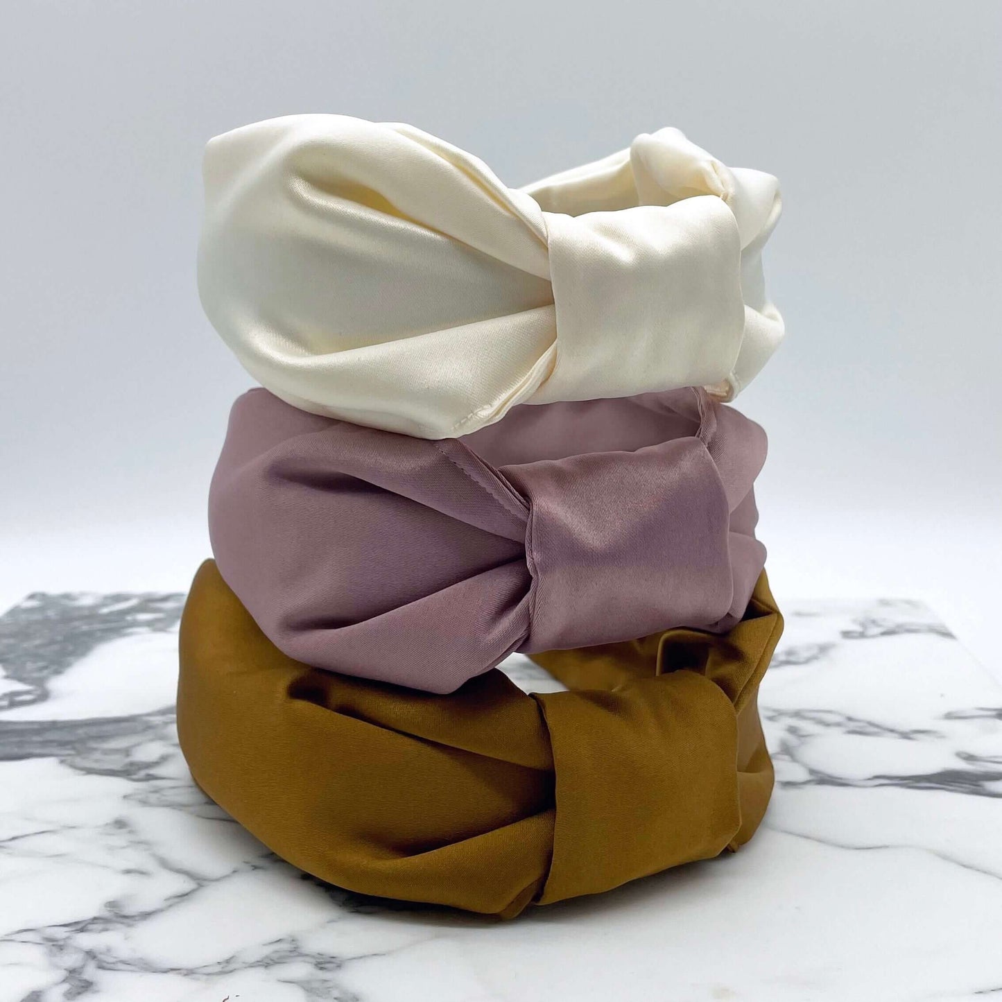 Three satin knotted headbands in a bow style, stacked. Pink, cream and gold.