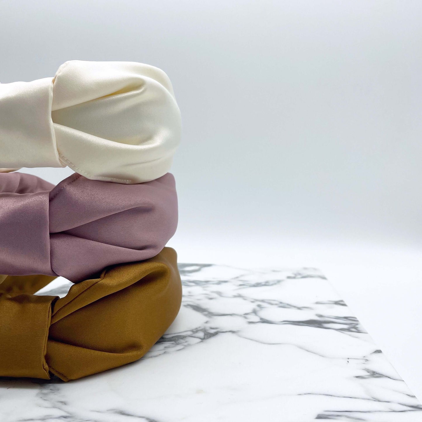 Three satin knotted headbands in a bow style, stacked. Pink, cream and gold.