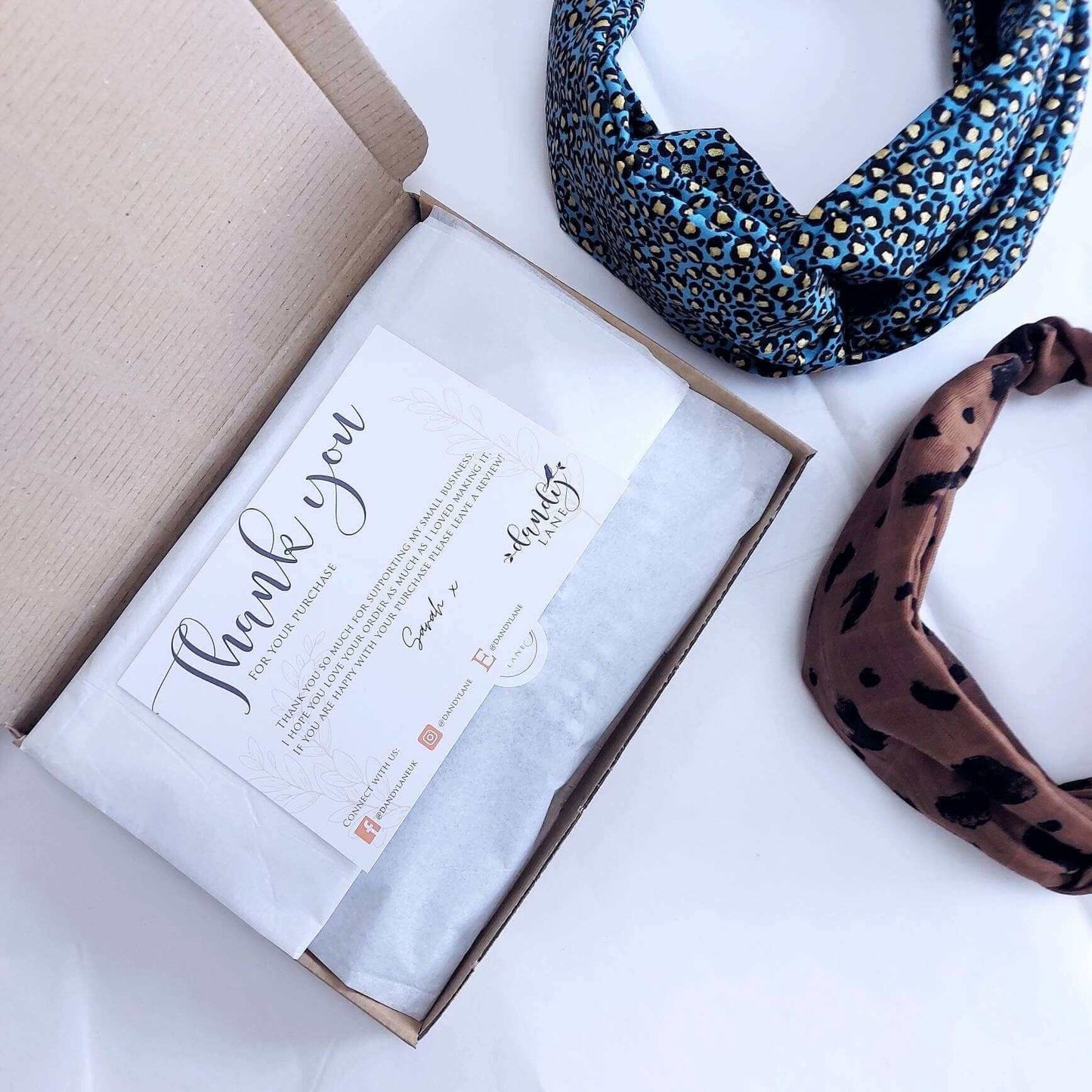 Two elasticated headbands next to a brown cardboard gift box with tissue paper and a thank you postcard