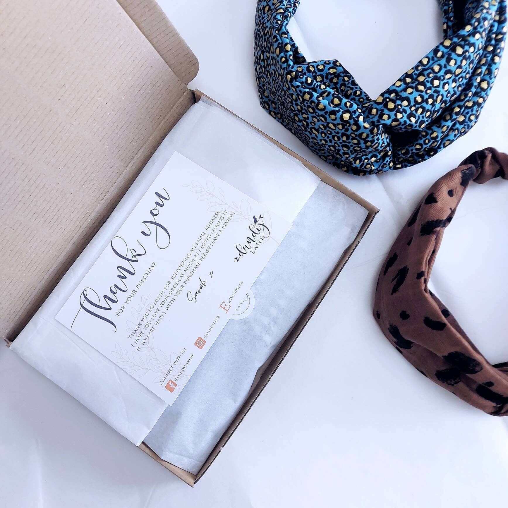 Two elasticated headbands next to a brown cardboard gift box with tissue paper and a thank you postcard