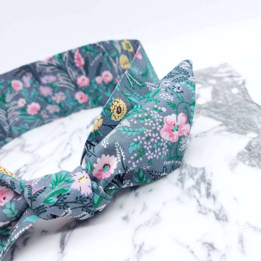 A summery, pale grey, ditsy floral cotton headscarf with tiny pink and green flowers, tied in a pretty knot.
