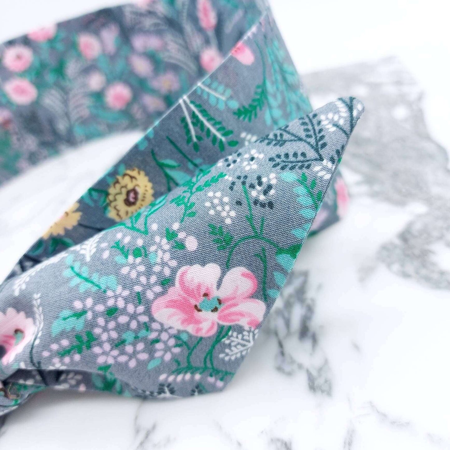 A summery, pale grey, ditsy floral cotton headscarf with tiny pink and green flowers, tied in a pretty knot..