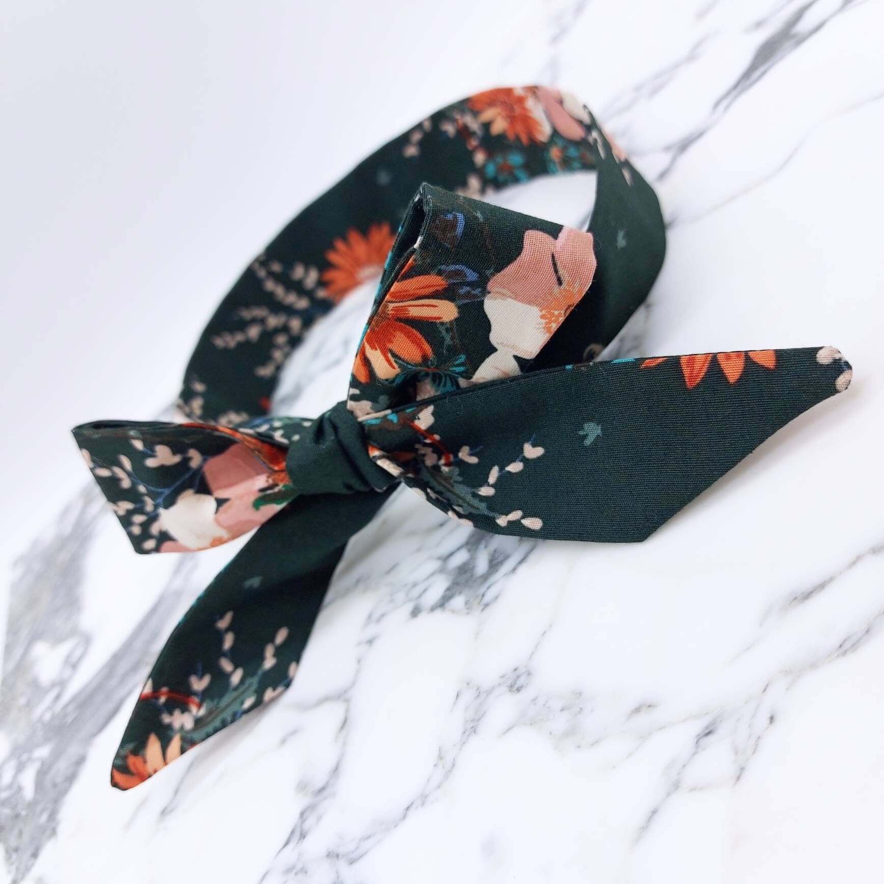 A dark green, floral cotton headscarf with pale pink, blue and orange flowers, tied in a pretty bow.