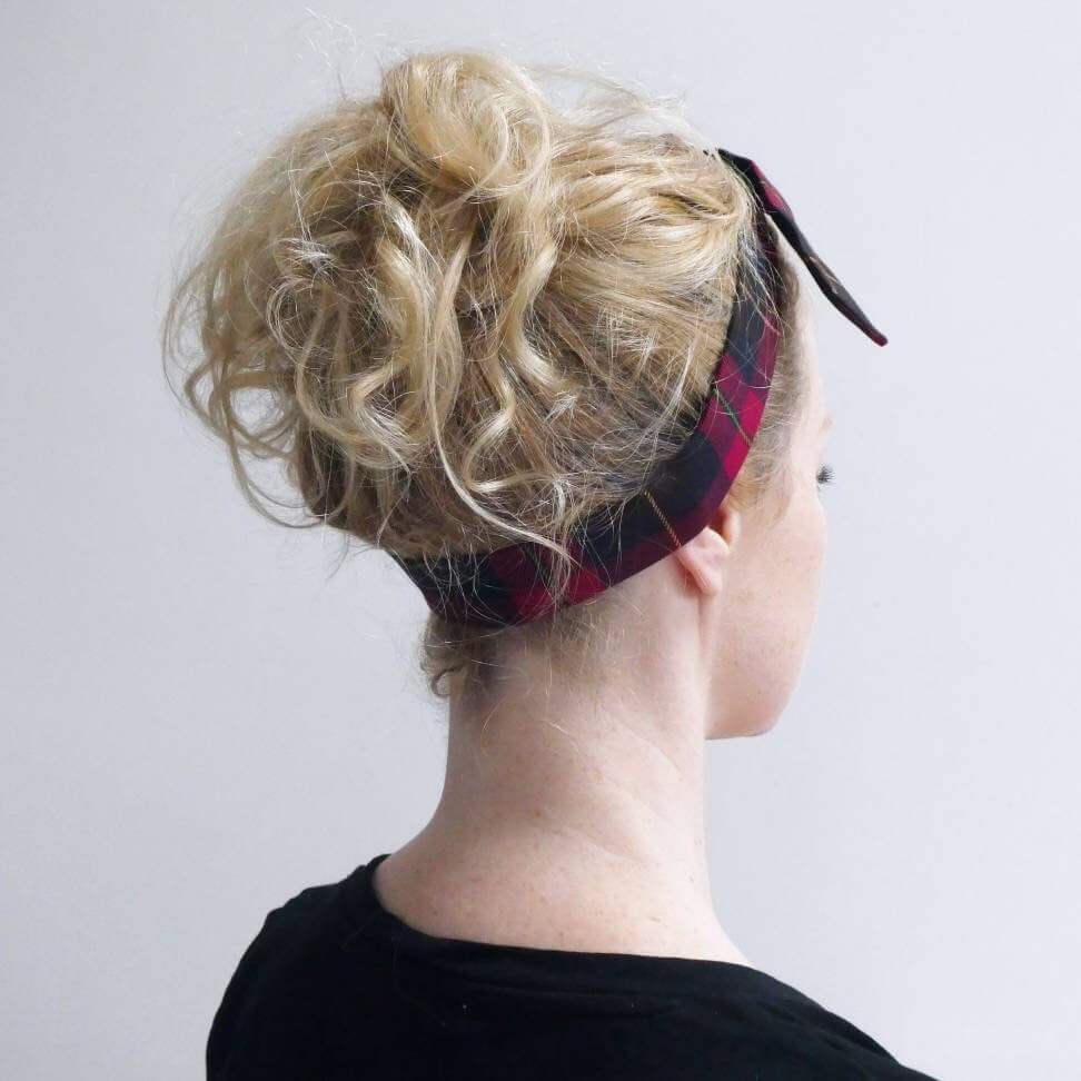 Model wears a dark cranberry and brown fabric, tartan paid tie headscarf, tied in a pretty knot.