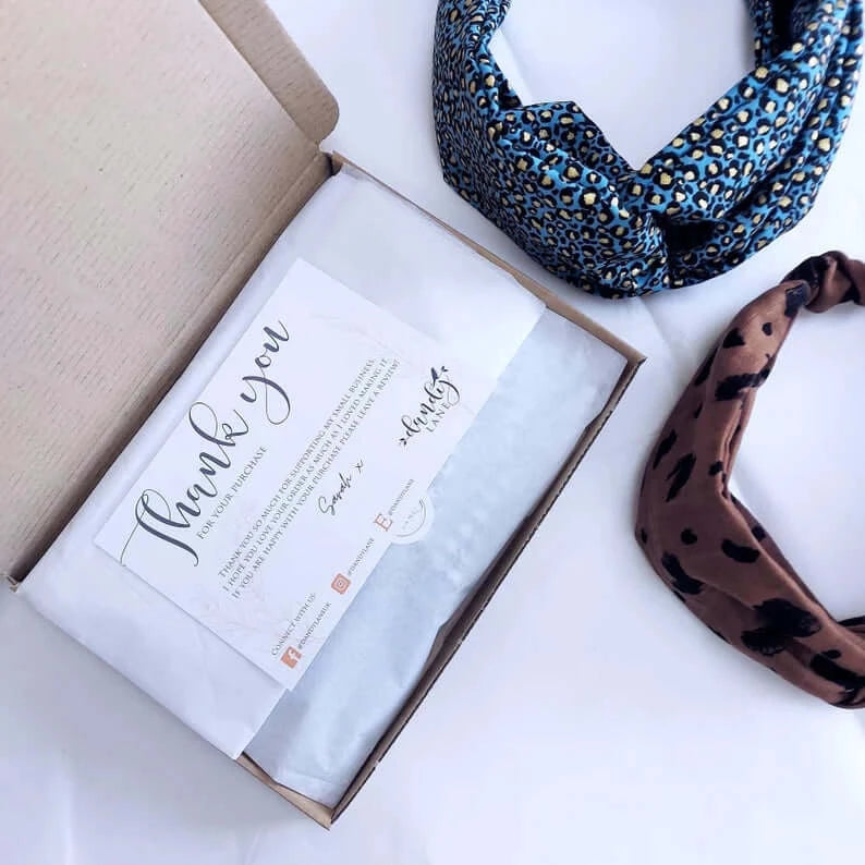 Two turban twist headbands next to a brown gift box, open with white tissue paper inside and a 'thank you' postcard on top.