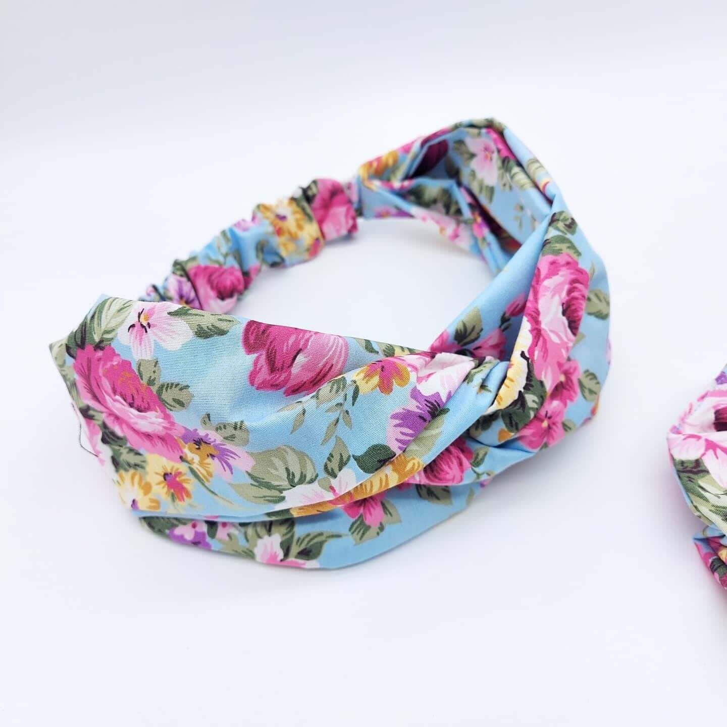 A pale blue, rose print floral cotton, turban twist headband with an elasticated panel at the back.