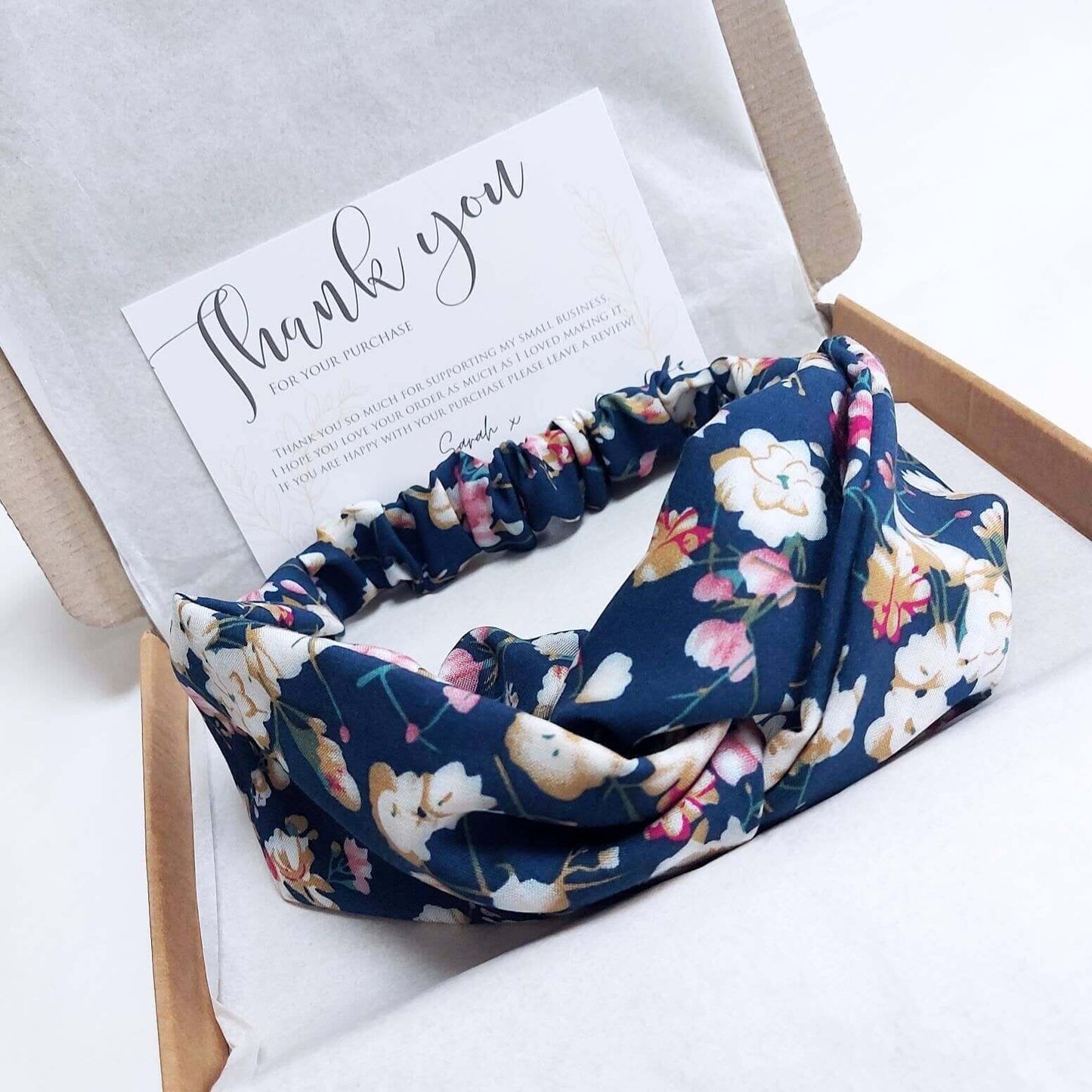 A soft, navy floral, turban twist headband with pops of pink, cream and yellow packaged in a brown gift box with tissue paper.