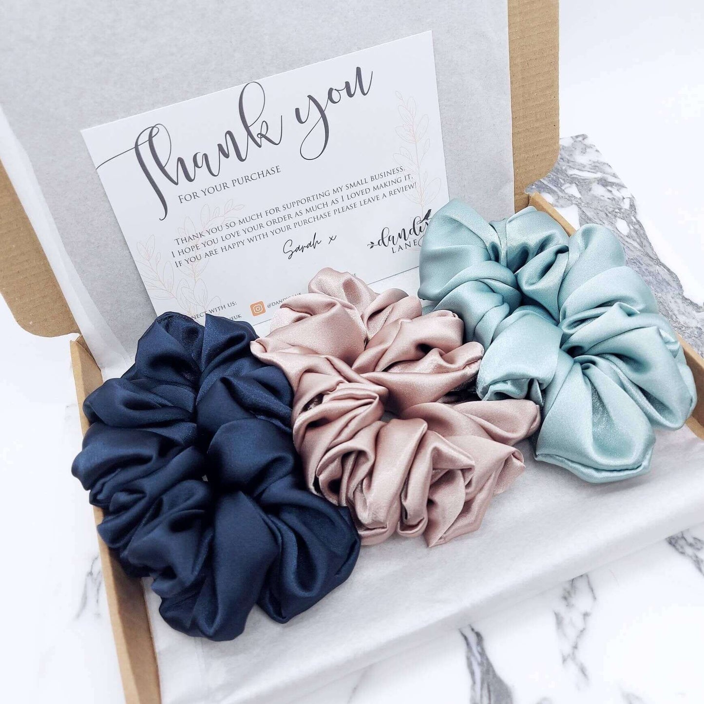 A tissue lined, brown gift box with three luxury, large satin hair scrunchies in midnight blue, rose and aqua.