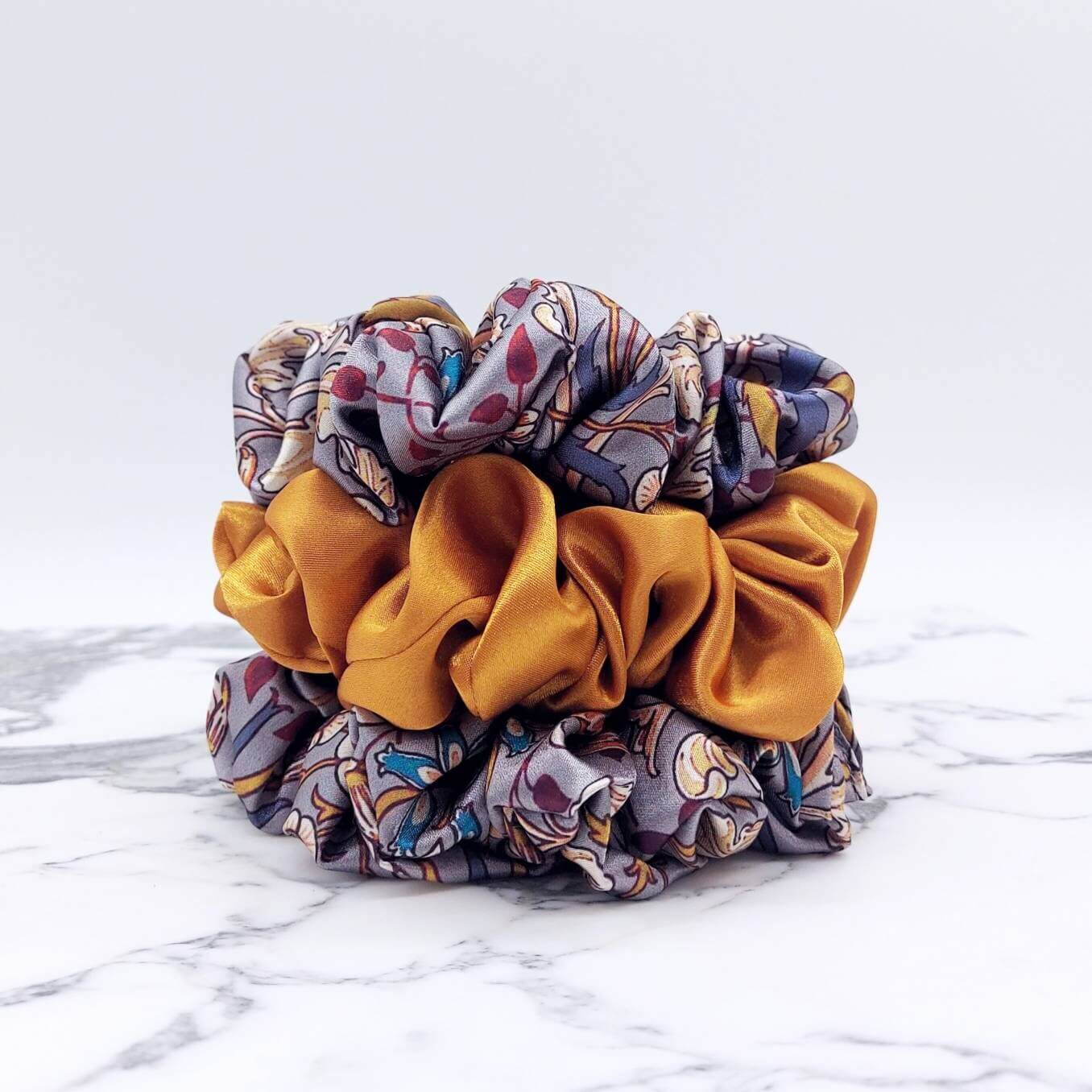 Three luxury satin hair scrunchies in a stack. Two grey patterned scrunchies with a gold satin scrunchie in the middle.