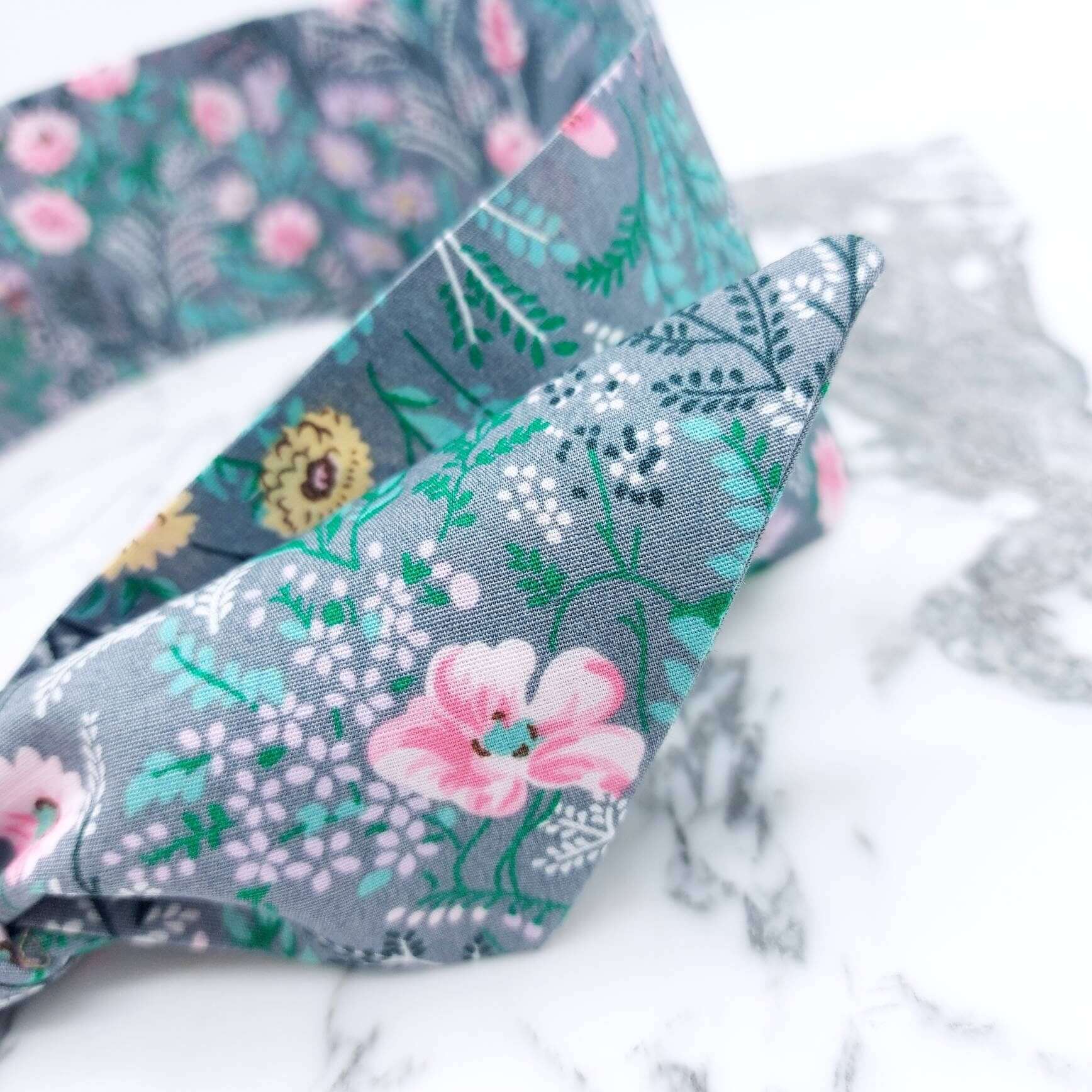 A summery, pale grey, ditsy floral cotton headscarf with tiny pink and green flowers, tied in a pretty knot..