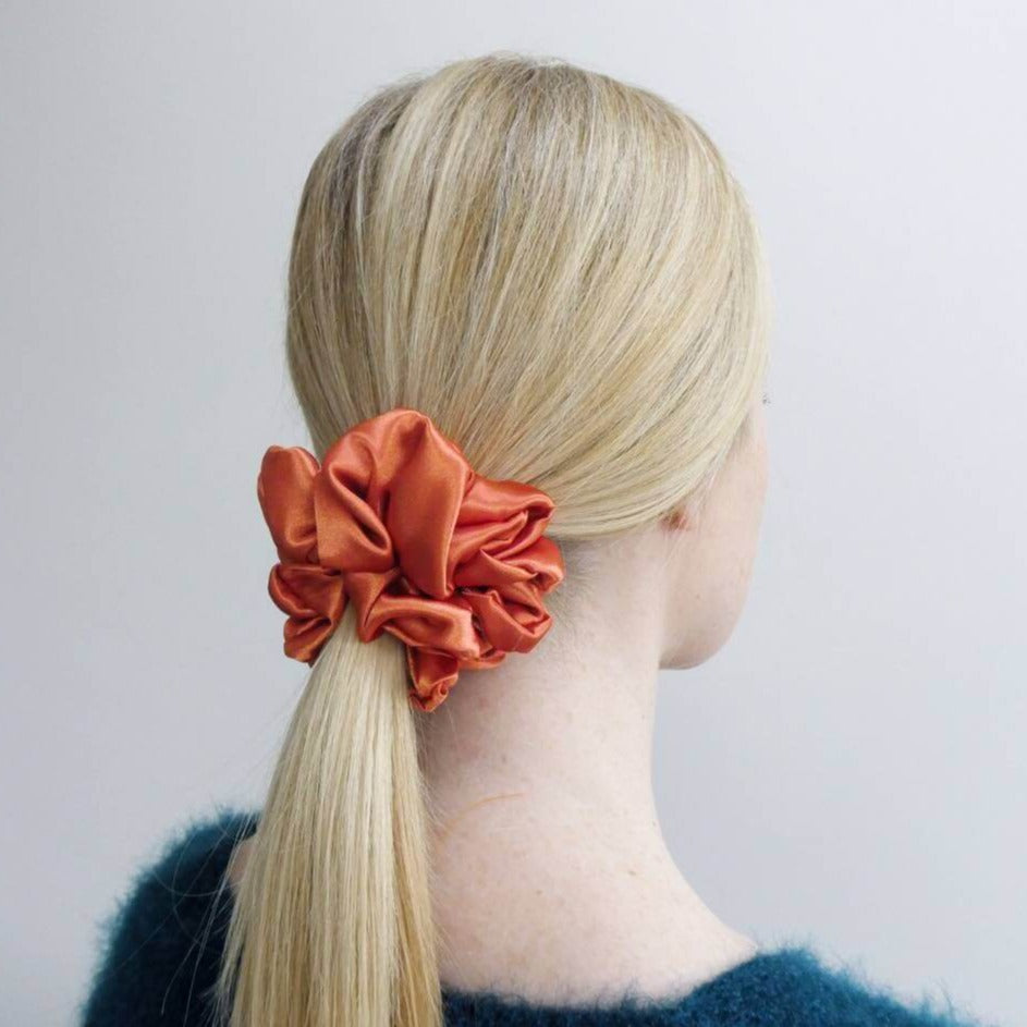 Model wears hair in a low ponytail with a large, luxury, tangerine, satin hair scrunchie tied around.
