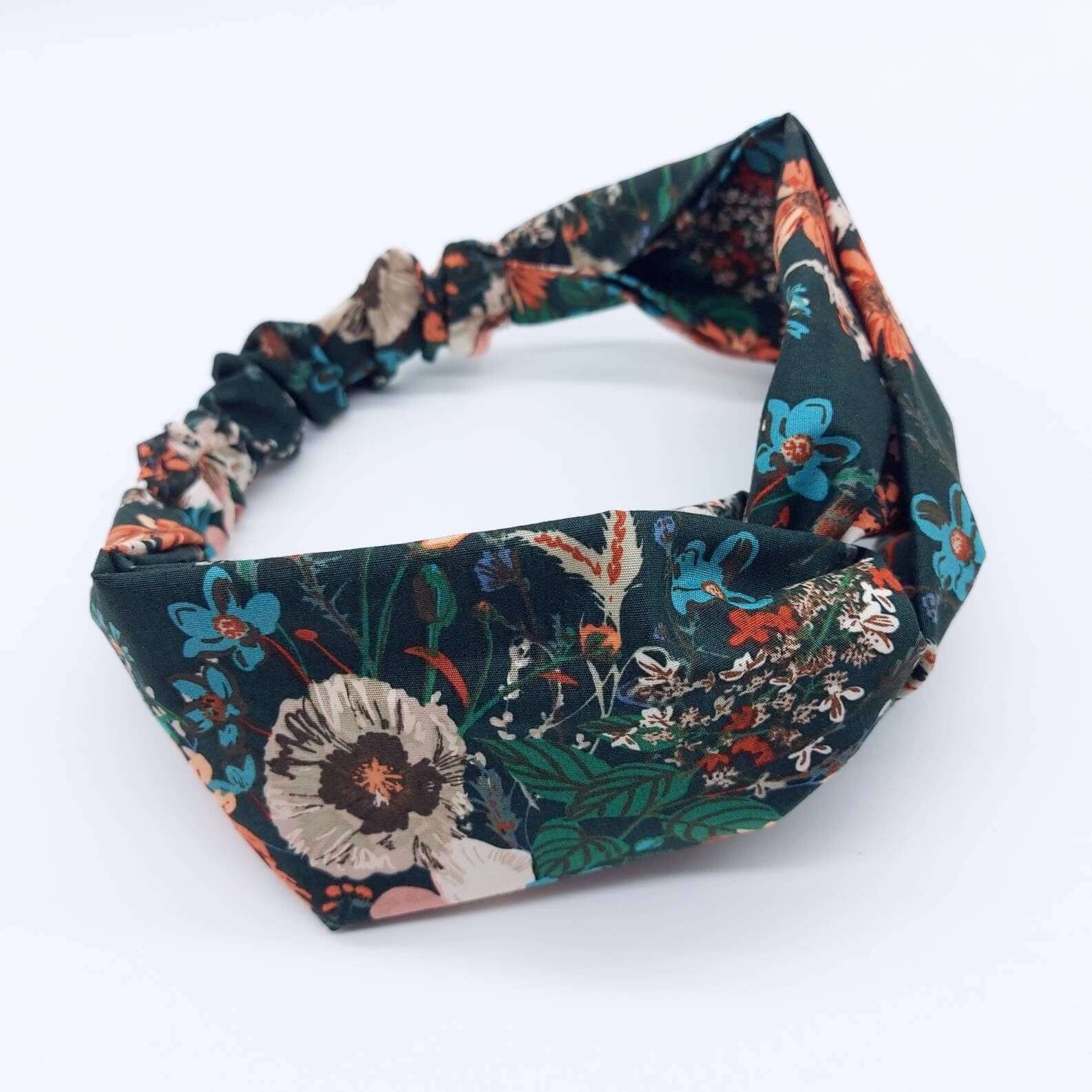 A dark green, orange and teal floral print, elasticated cotton headband, with a turban twist design at the front.