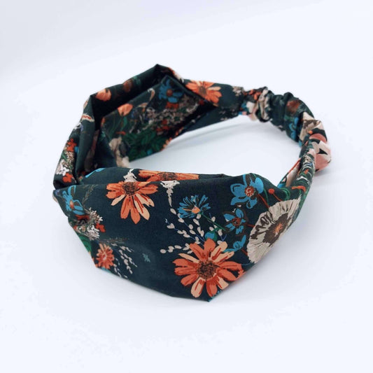 A dark green, orange and teal floral print, elasticated cotton headband, with a turban twist design at the front.