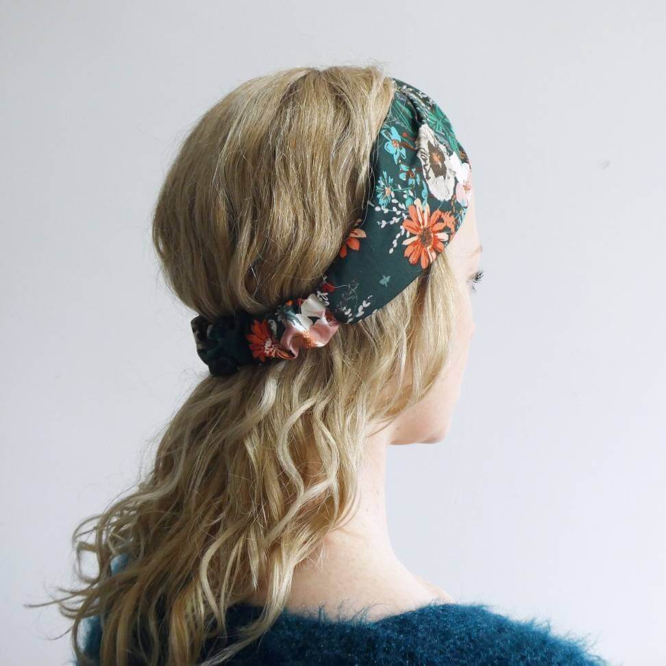 Model wears a dark green, orange and teal floral print, elasticated cotton headband, with a turban twist design at the front.