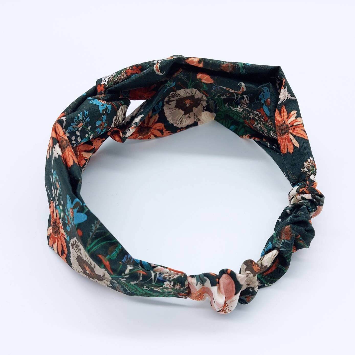 A dark green, orange and teal floral print, elasticated cotton headband, with a turban twist design at the front.