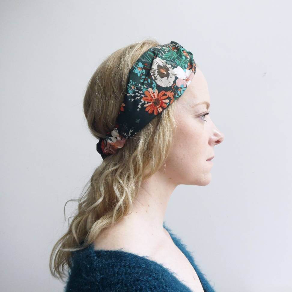 Model wears a dark green, orange and teal floral print, elasticated cotton headband, with a turban twist design at the front.