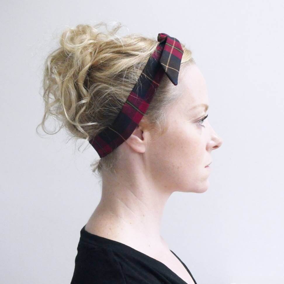 Model wears a dark cranberry and brown fabric, tartan paid tie headscarf, tied in a pretty knot.