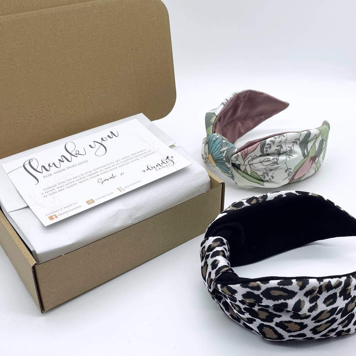 Two knotted style headbands next to a brown cardboard gift box with tissue paper and a thank you postcard inside.