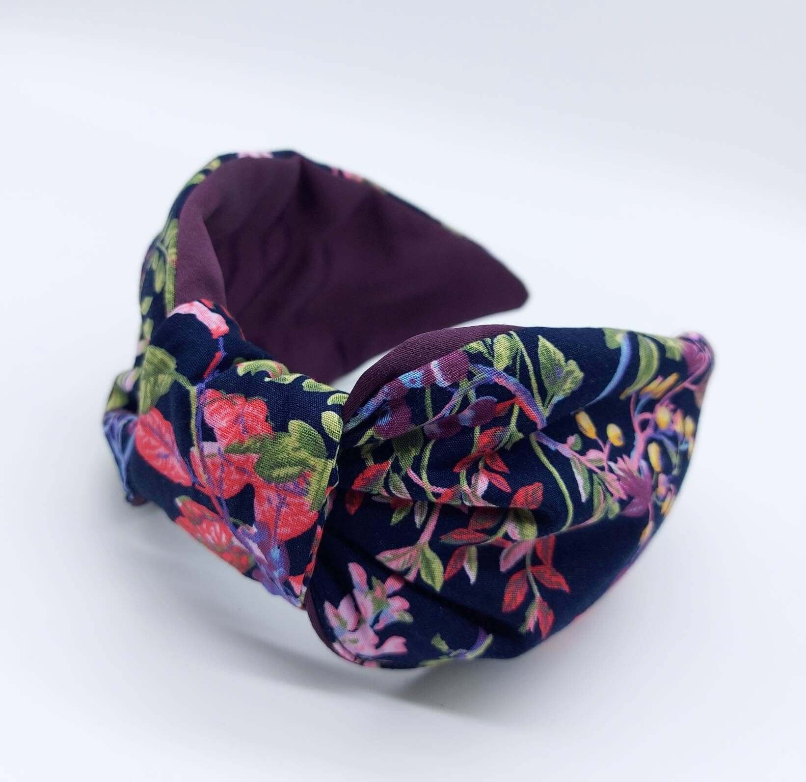 A bow-style, navy blue, red, purple and green floral fabric knot headband with purple satin lining underneath.