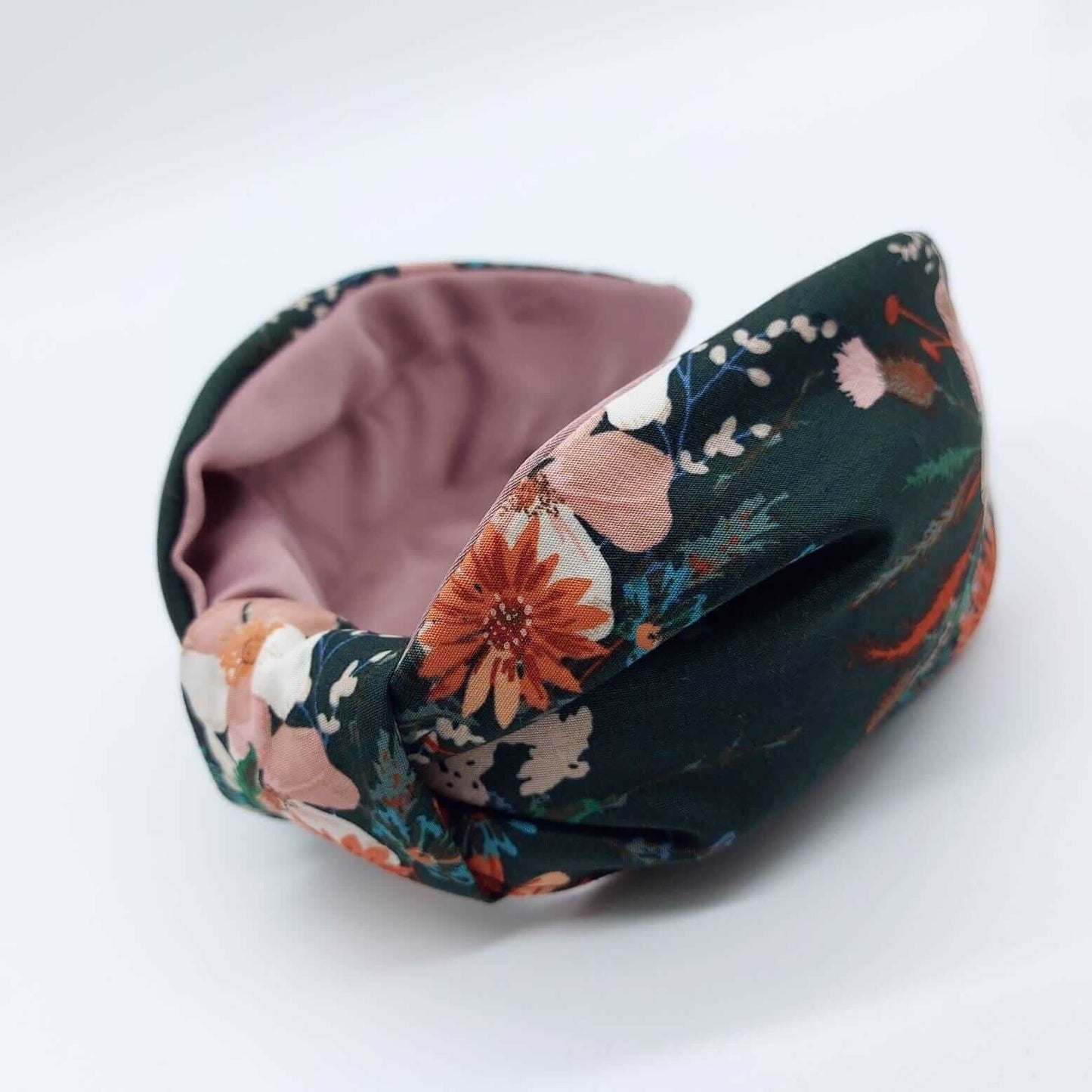 A bow-style, knot headband made from dark green fabric with orange and pink flowers and a pink satin lining underneath.