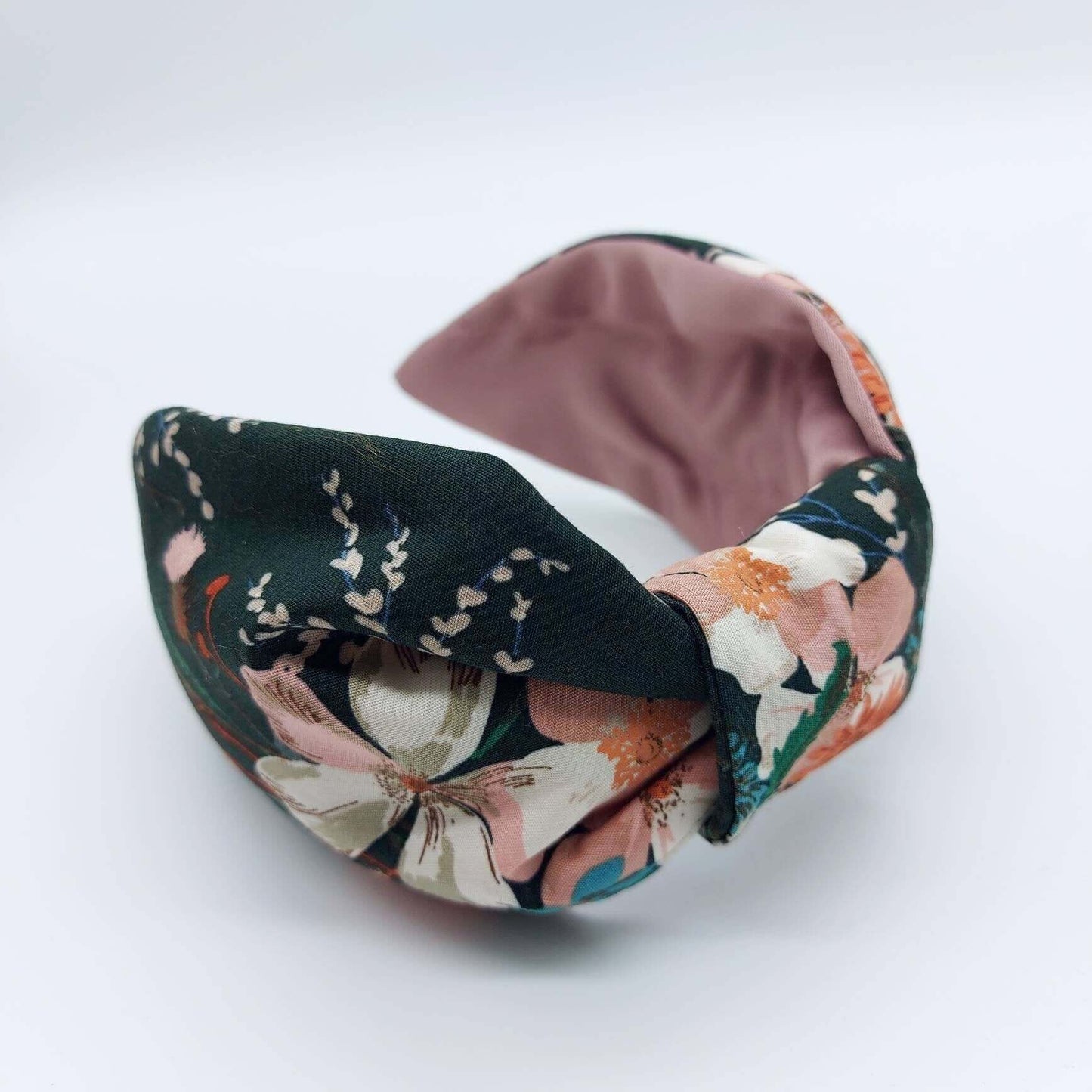 A bow-style, knot headband made from dark green fabric with orange and pink flowers and a pink satin lining underneath.